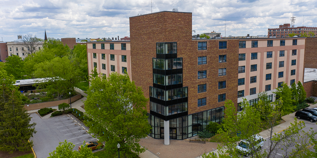 A brick building with glass windows. A parking lot is to the left and green trees are in the front.