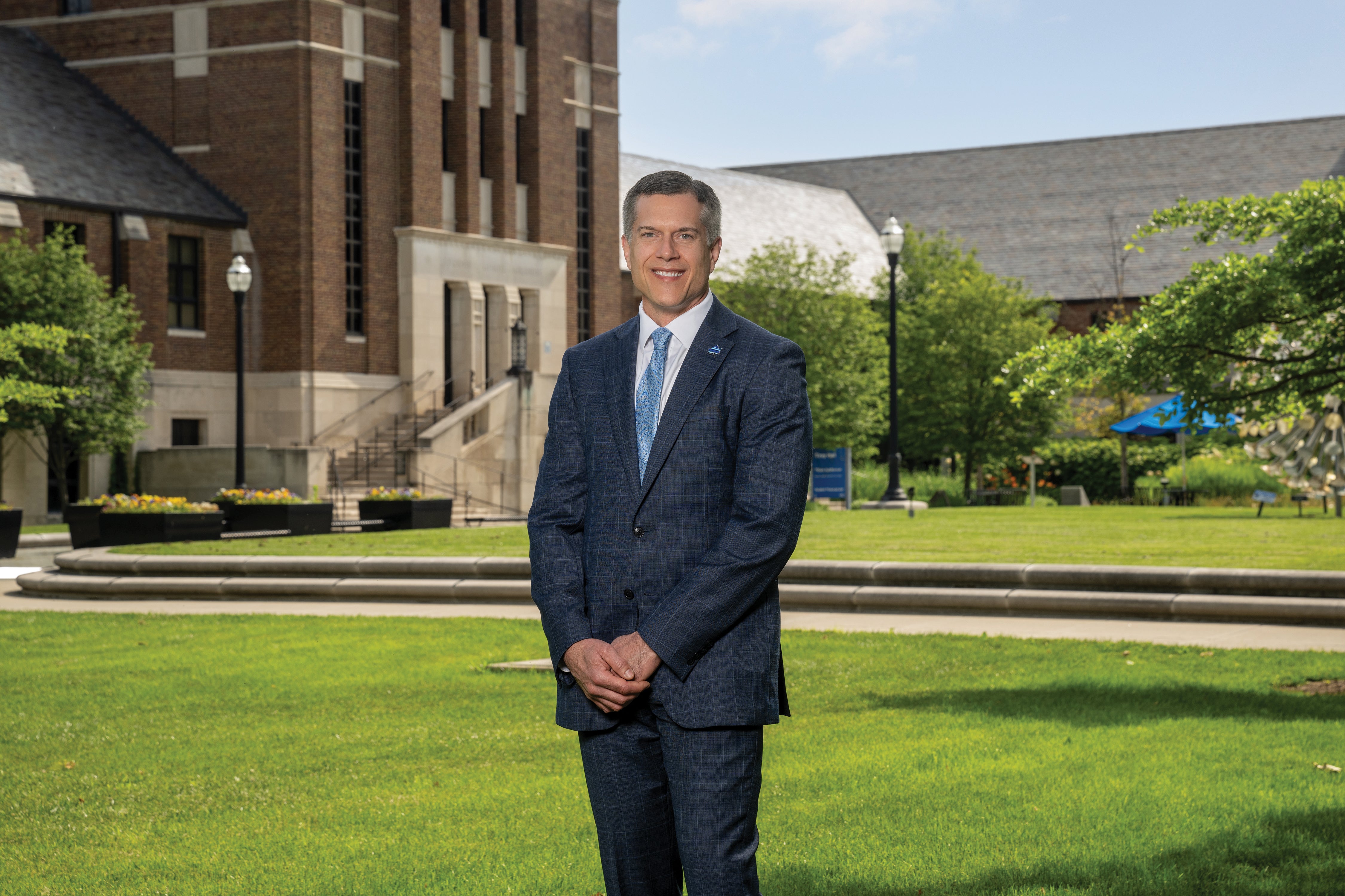 Indiana State University President Mike Godard poses outside with his hands folded. He is a white middle-aged male with greying hair. He wears a dark blue dress suit, a white undershirt, and a light blue tie. Visible behind him is a brick building, a grassy lawn, and other greenery.