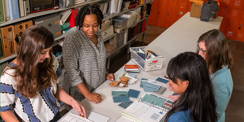 A female design professor stands alongside three female students as they review color and design swatches for a class project. The narrow worktable has color samples – blue, gray, mauve – spread across it.