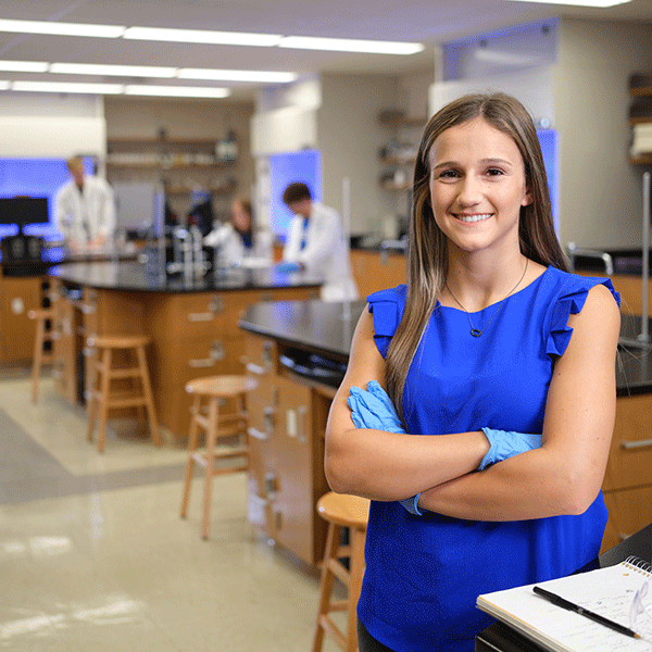 A female student with brown hair and wearing a sleeveless blue dress, her arms crossed and with blue latex gloves on her hands, stands in the foreground with a science laboratory visible behind her. People in white lab coats are visible working in the background.