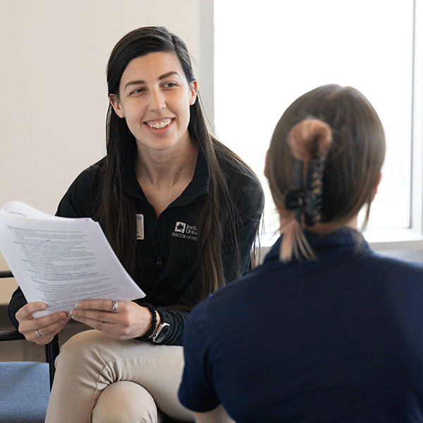 A smiling Indiana State student with long dark hair talks with another woman who is only visible from behind. The student is wearing a black Indiana State shirt and tan pants. Her legs are crossed and she is holding stapled papers in her hands.