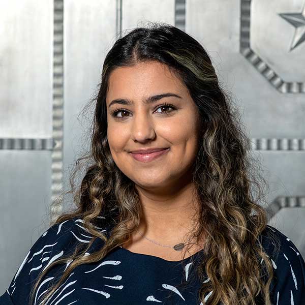 A woman with tanned skin and dark brown hair with light brown highlights poses in front of a silver wall. She wears a navy blue and light blue dress shirt.