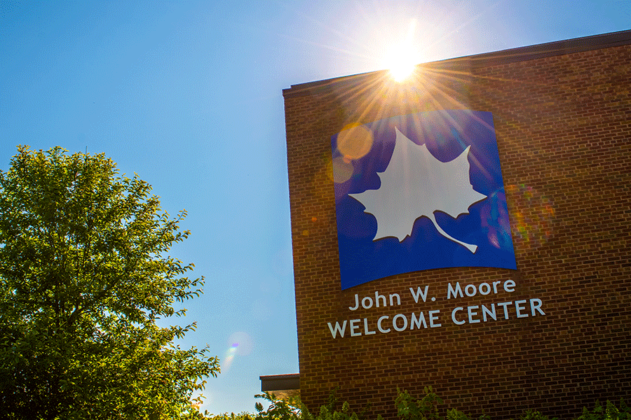 The sun is partially visible over the red brick wall of the John W. Moore Welcome Center. Rays of light shine over top the large white Sycamore leaf on the blue background that appears on the side of the building with the name underneath. A green tree is also visible to the right against the blue sky.