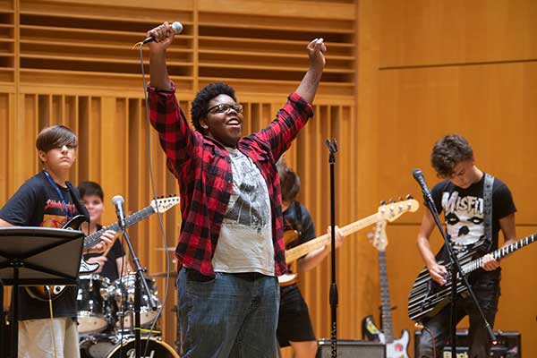 Five young men of mixed races perform music. In the front of the group is a Black man wearing a white T-shirt, a black-and-red plaid jacket, and blue jeans. He holds a microphone with his arms raised. Behind him are four males wearing black T-shirts. Three hold guitars and one sits behind a drum set.