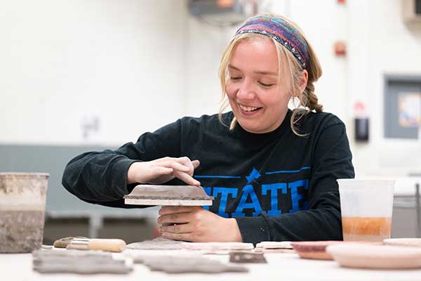 A young, smiling blonde woman, wearing a multicolored scarf in her hair and a navy blue sweatshirt that says STATE in royal blue letters, rubs her finger along the top of an unfinished clay disc on a potter’s wheel. Other flat clay items are visible on the table in front of her, and a bucket of clay can be seen to her right.