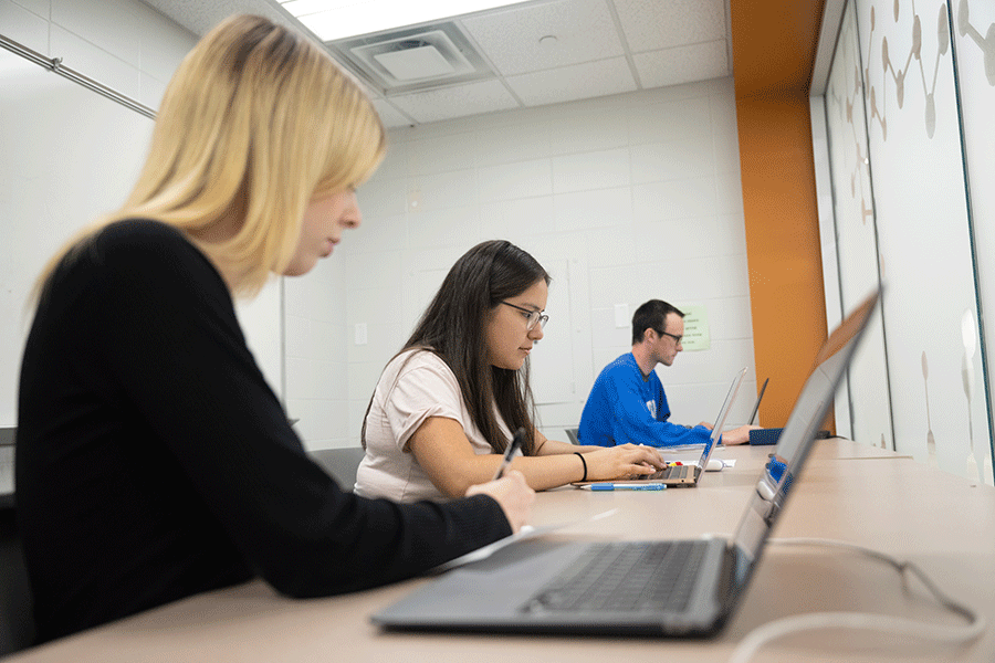Three students – a blonde woman in a black sweater, a woman with brown hair and wearing a white shirt, and a man with dark hair and glasses wearing a blue sweatshirt, type on their laptops in a well-lit, white-walled classroom.