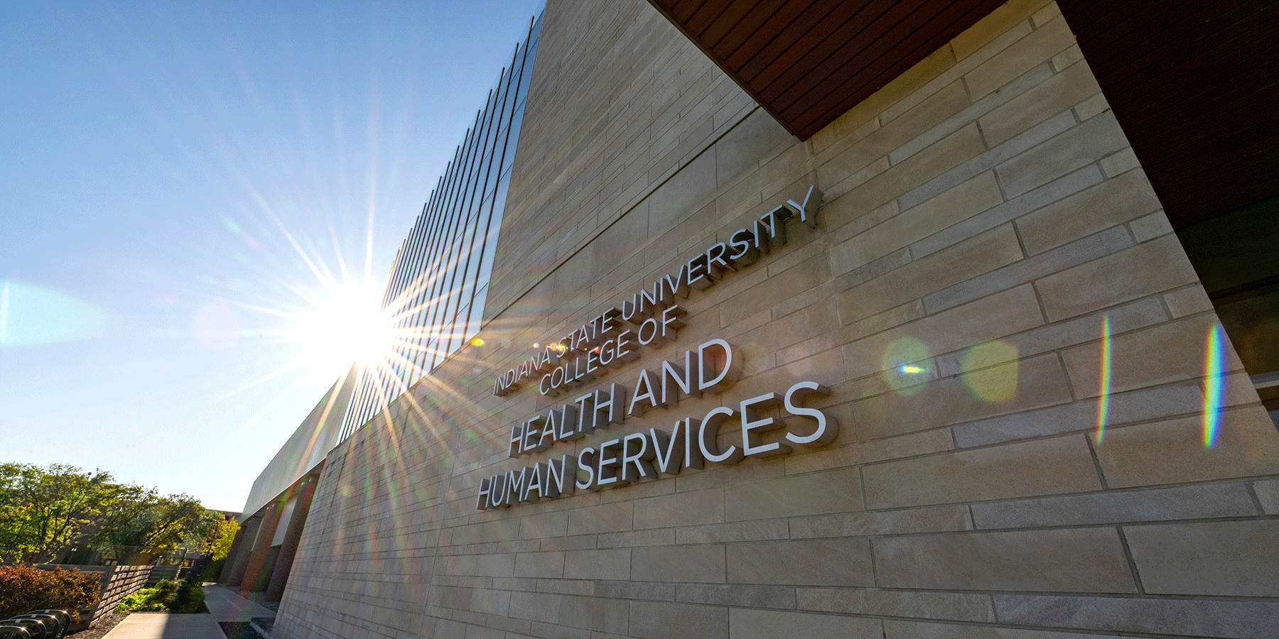 An exterior view of the College of Health and Human Services building, a brick building with glass windows on the left side of the building. “Indiana State University College of Health and Human Services” lettering is on the front side. Green trees are to the left of the building.