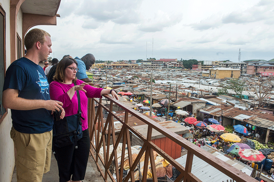 Adult male in navy Indiana State University t-shirt stands next to shorter adult woman in pink top on balcony overlooking a city in another country.