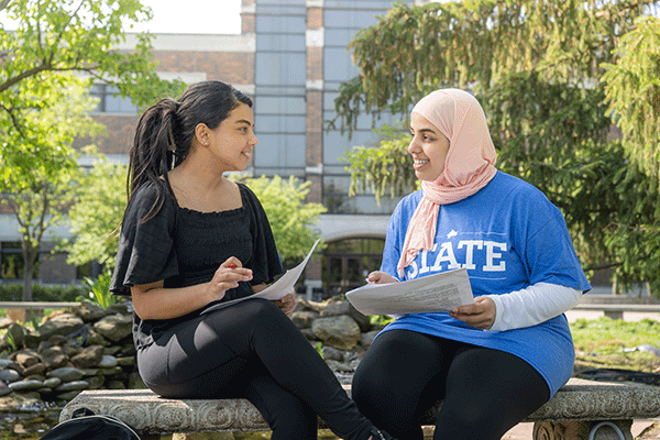 Two females sit on a bench talking while holding papers