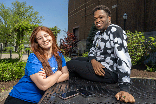 Two students, one female sitting at a table and one male sitting on the table, smile and pose