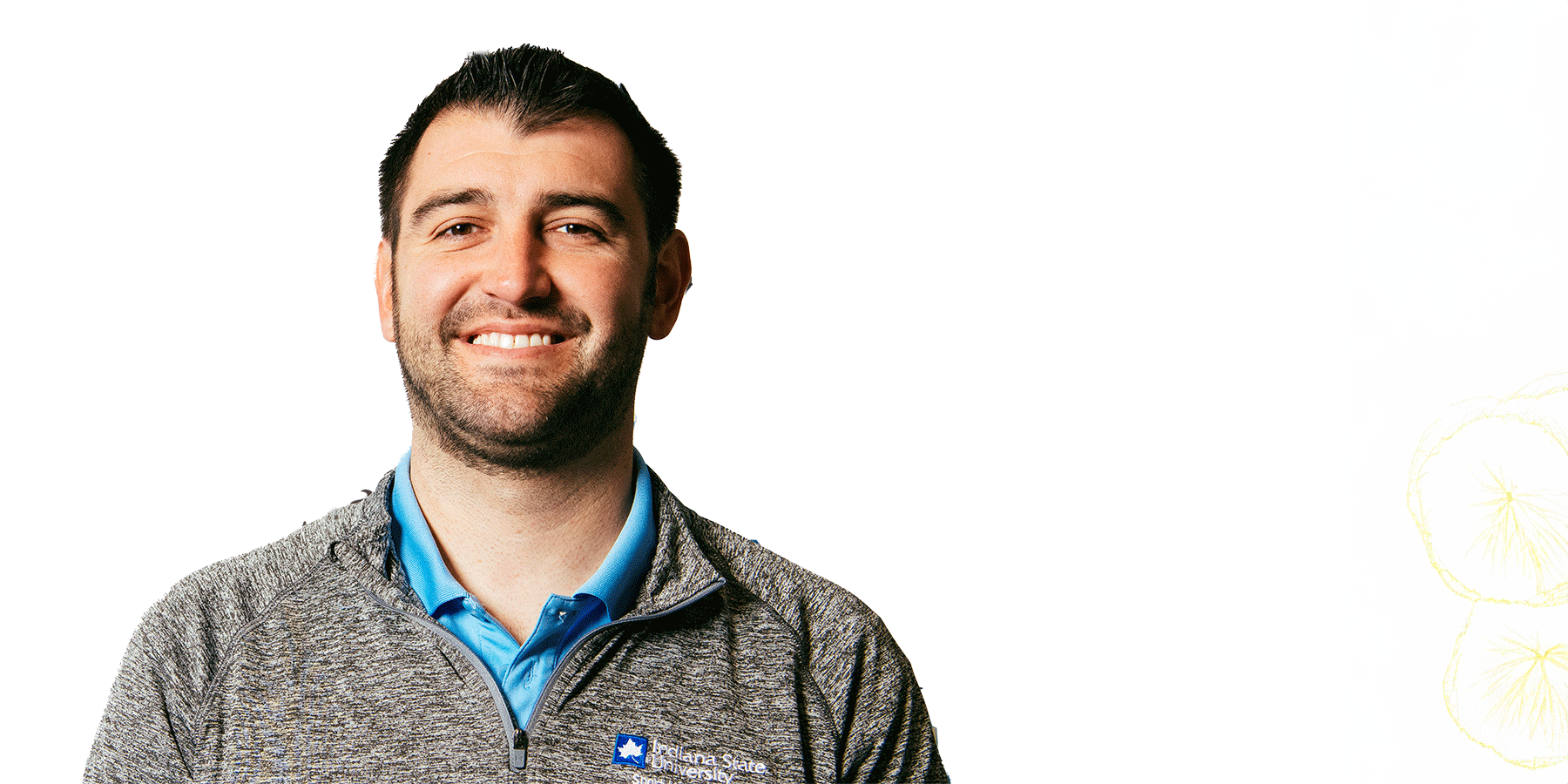 A white male with short black hair with a matching mustache and beard poses before a white backdrop. He wears a grey Indiana State University zipped-up jacket with a blue undershirt.