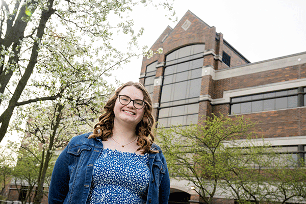 A photo of a white female student with brown hair and glasses, who stands at a distance in front of a tall brick building with many glass windows. It is the John T. Myers Technology Center. The woman is wearing a blue dress with a white pattern on it and a blue jean jacket. Trees with white flowers and dark branches are visible in the background.