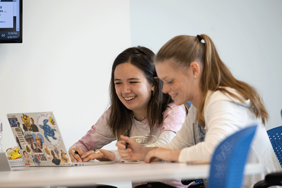 Two young white female students sit at a table and laugh together. One is staring at her laptop. She has shoulder-length black hair and wears a pink and white top. The other is staring at a paper on the table. She has brown hair in a ponytail and wears a white top. A white wall forms the background.