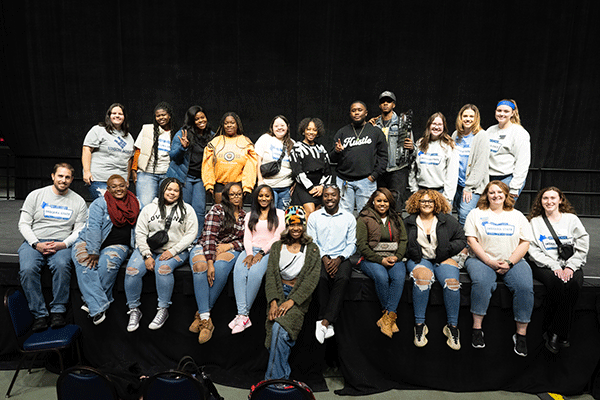 A large group of students gather along the edge of a stand and pose for a photo following a student organized university event.  