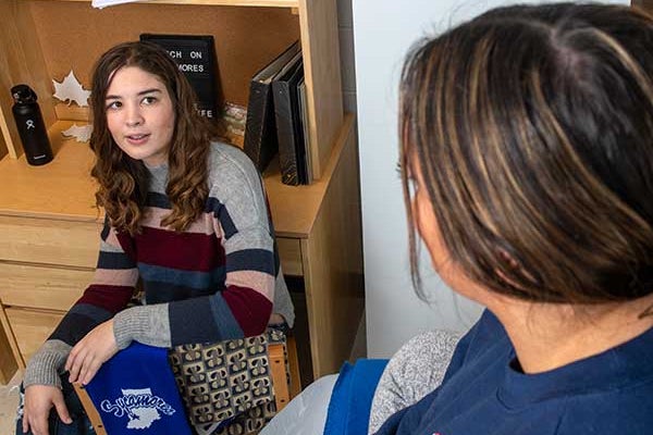 A student seated on a chair in her residence smiles up at her roommate, seated on the bed. An Indiana State University scarf is visible draped over the chair arm.
