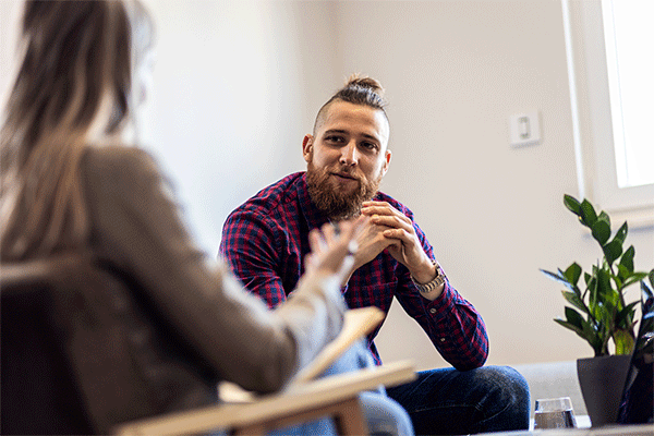 A white male with a beard and hair in a bun and facing the camera listens as a counselor in the foreground with her back to the camera and slightly out of focus speaks. 