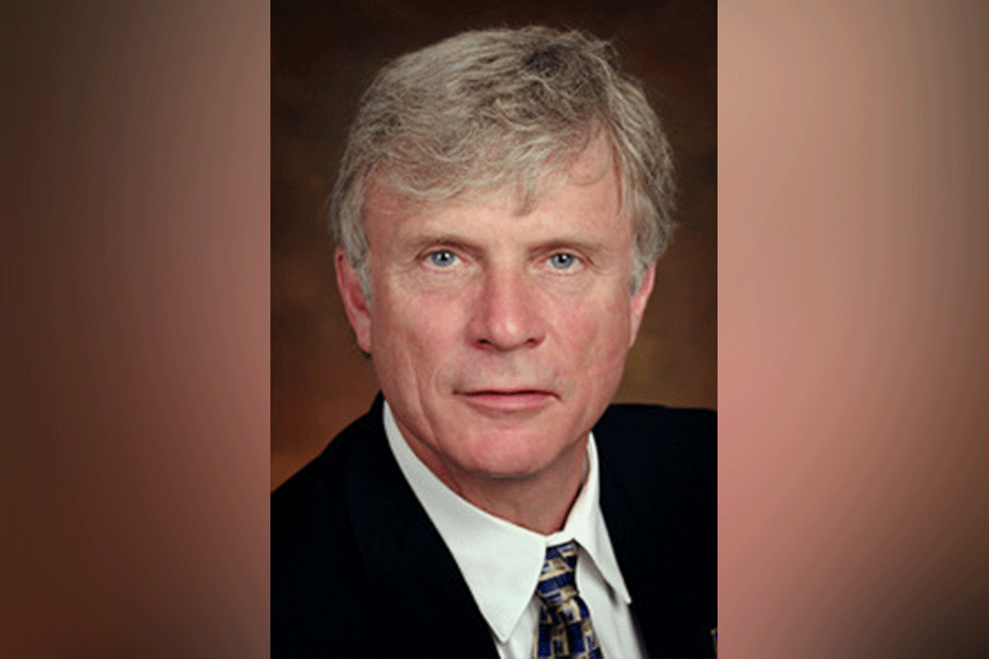 A man (President Lloyd W. Benjamin III) with short greying hair. He looks straight ahead. He wears a black suit with a white shirt and a tie. A light brown border is around the photograph.&nbsp;