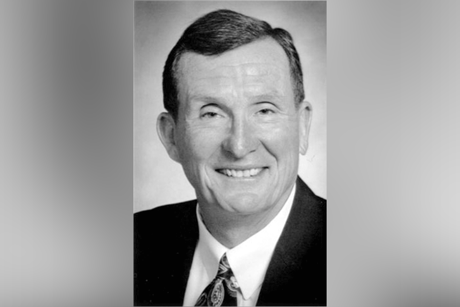 A black-and-white photograph of a man (President John William Moore) with short hair. He smiles as he looks straight ahead. He wears a suit with a white shirt and tie. A grey border is around the photograph.&nbsp;