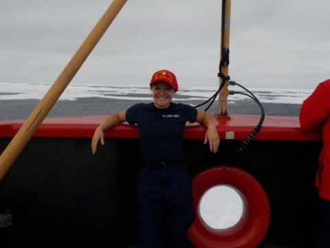 Person in a red cap and navy shirt standing on a boat deck, smiling with arms resting on the railing; a red life ring is mounted beside them, and icy ocean waters stretch into the background.