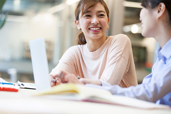 Two smiling female students of Asian heritage sit in a well-lit study area talking and laughing. The woman at left faces the camera and looks at her companion. She has medium-length brown hair in a ponytail and wears a light beige, long-sleeved shirt. Her friend has black hair and is looking away from the camera at the edge of the frame. She wears a blue-and-white pin-striped shirt, holds a pen, and has a laptop open before her. Books, pens, and notepads are visible but out of focus on a table.