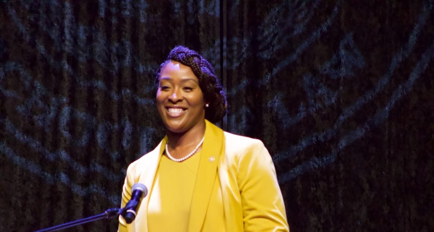 A Black woman stands in front of a microphone, wearing a yellow blazer and matching yellow shirt, accessorized with a pearl necklace. The background is dark with indistinct patterns.