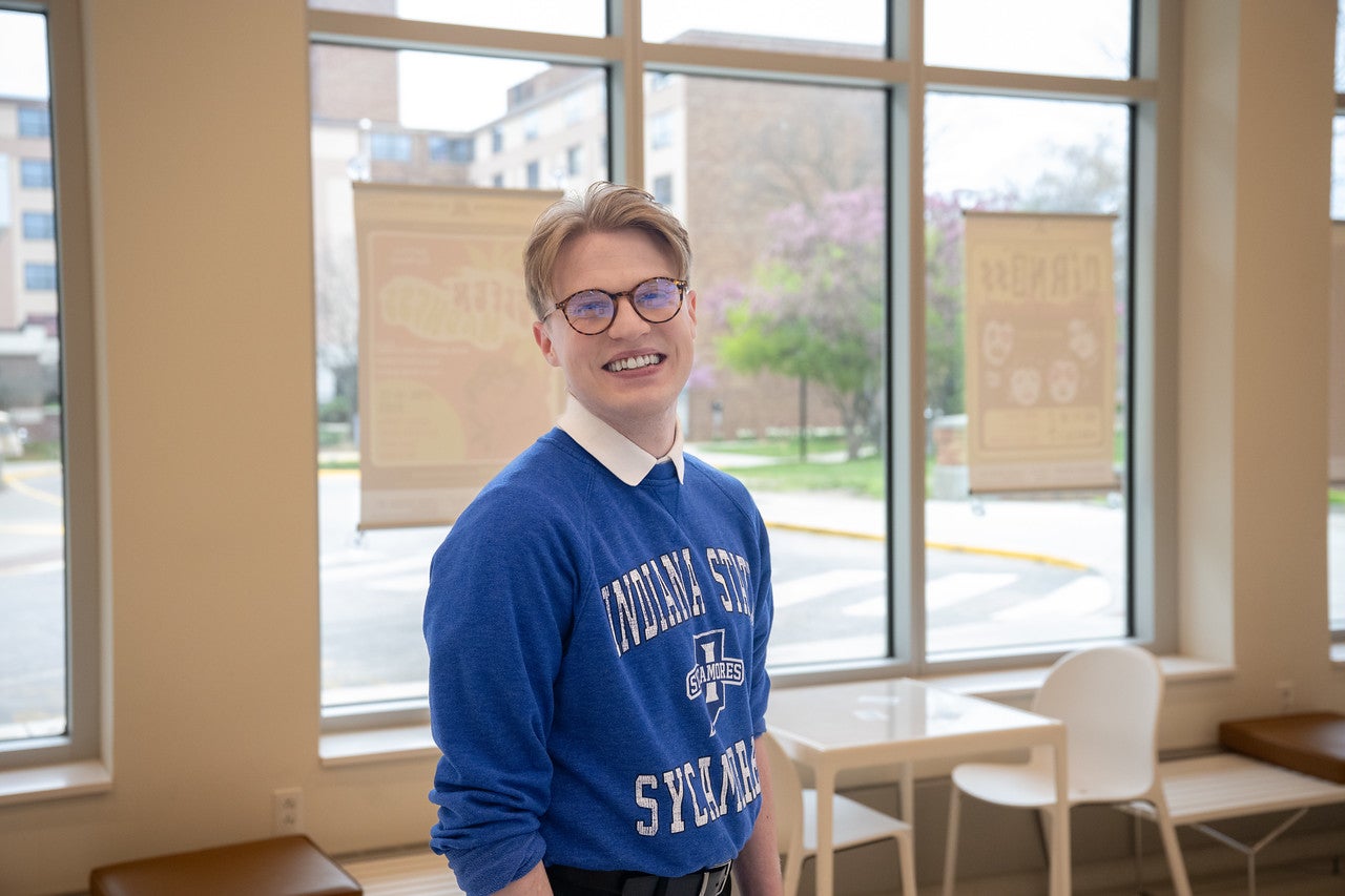 A white male with short blond hair is wearing a blue "Indiana State Sycamores" sweatshirt. He stands indoors in front of large windows. Outside the windows, trees and buildings are visible. Inside, there are chairs and tables near the windows.