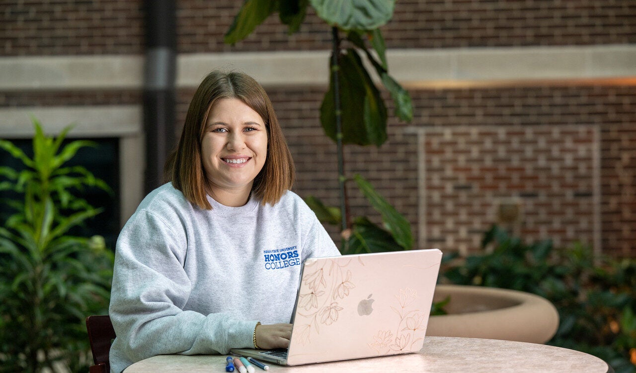 A white female with shoulder-length brown hair is sitting at a table using an Apple laptop. She is wearing a gray sweatshirt with "Honors College" written on it in blue text. Pens or markers are placed on the table next to the laptop. The background includes plants and a brick wall.