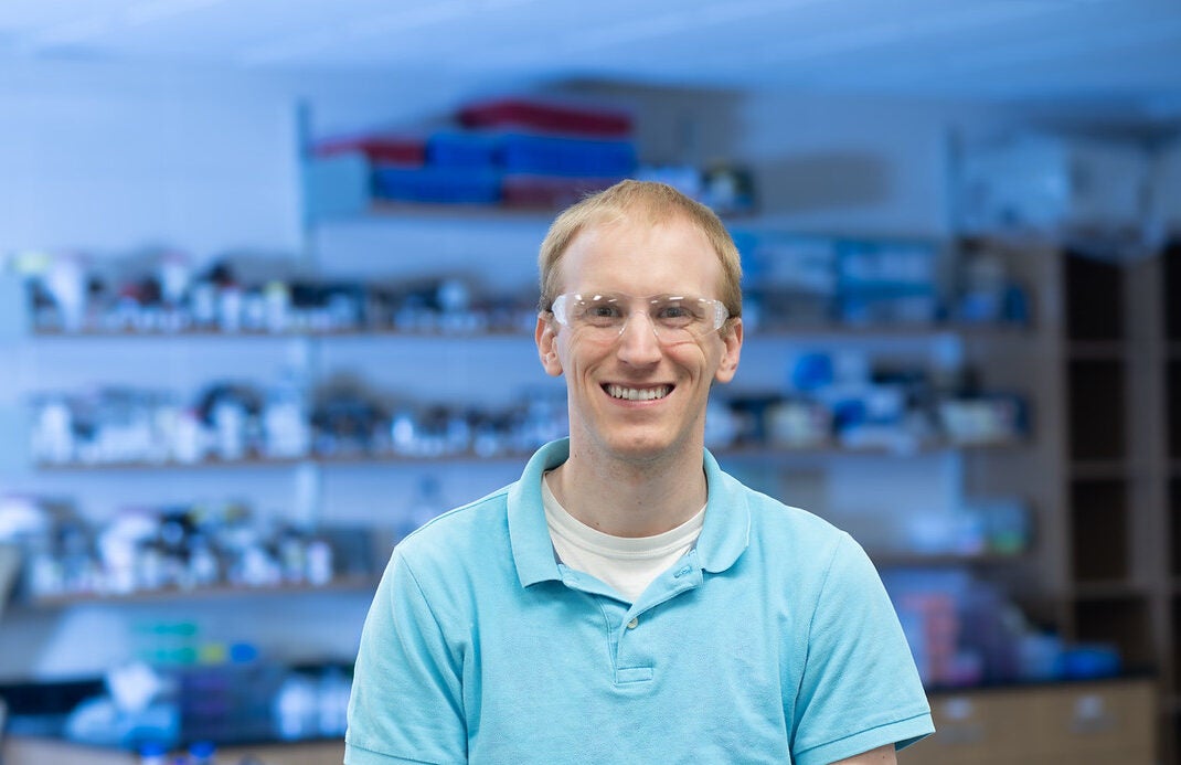 A white male with short blond hair stands in a laboratory setting. He wears a light blue polo shirt over a white undershirt and safety glasses. The background shows shelves filled with various laboratory equipment, containers, and supplies.