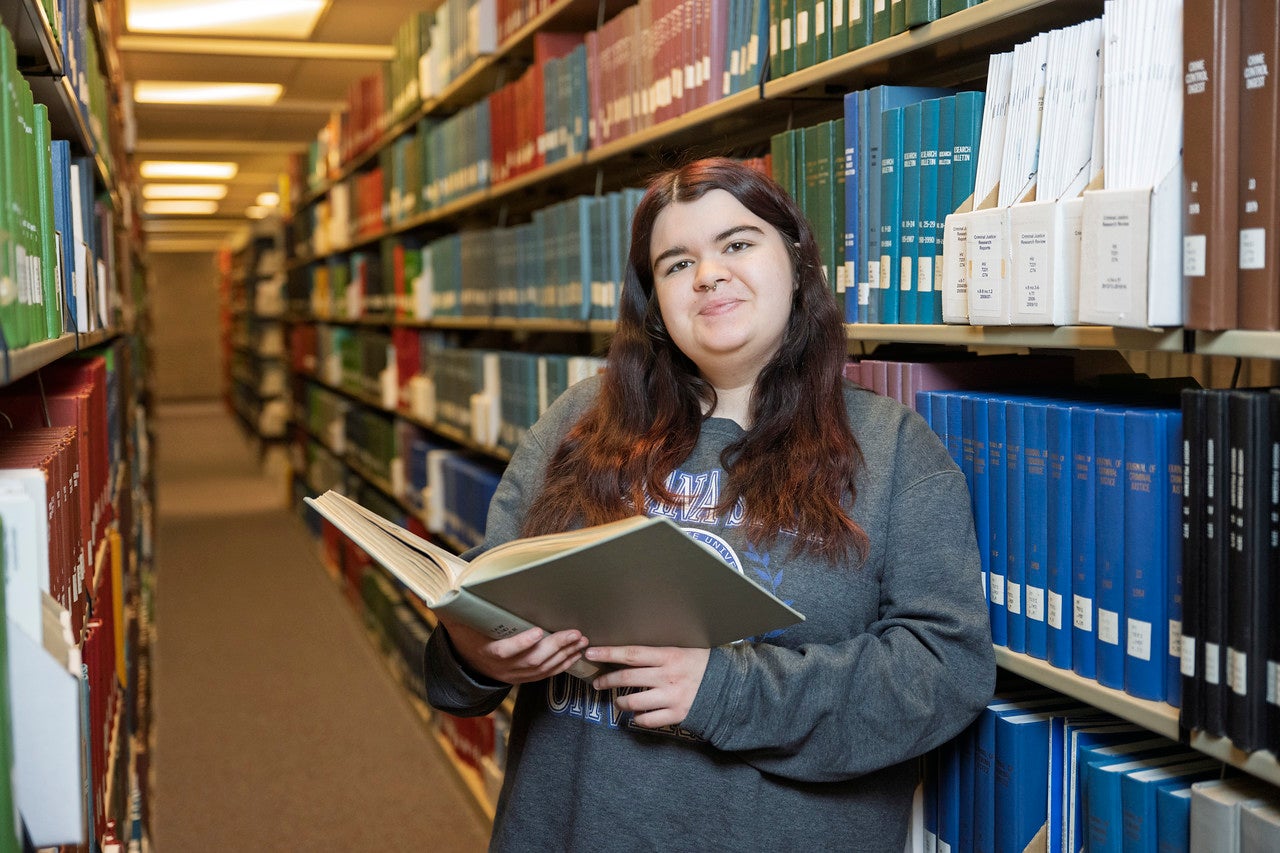 A white female with long brown hair, wearing a gray sweatshirt, is standing in a library aisle filled with bookshelves on both sides. She is holding an open book. The bookshelves are neatly organized with various colored books.