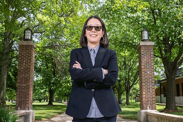 Dr. Katherine Lee stands outdoors on the Indiana State University campus, on a pathway between two brick pillars topped with lanterns. The background includes green trees and grass. Dr. Lee is wearing a dark blazer over a light-colored shirt and has her arms crossed.