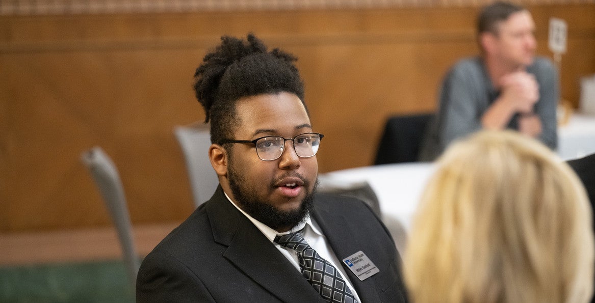 A Black male in a black suit with a patterned tie and a name tag is seated at a table in what appears to be a formal setting. The background shows another person sitting at a table. The back of a blonde-haired person's head is visible in the foreground.