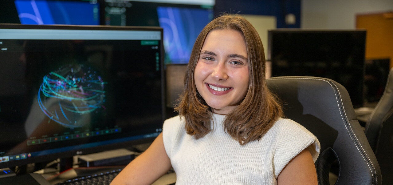 A white female with shoulder-length brown hair is sitting in front of a computer monitor displaying a colorful, abstract digital visualization. She is wearing a white sleeveless top and is seated in a gray office chair. Multiple monitors and computer equipment are visible in the background.