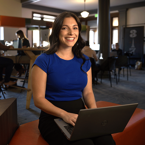 A female with long dark brown hair wearing a blue shirt and black pants sits on a dark orange bench in a modern, well-lit indoor space, possibly a café. A laptop is resting on her lap. Other people are visible in the background, working on laptops.