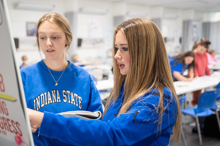 Two white female students wearing blue Indiana State sweatshirts are standing in a classroom. One person is pointing at something on a whiteboard while the other person is holding a notebook and looking at the whiteboard. Visible in the background, several other people are seated at desks. The classroom has white walls.
