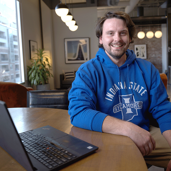 A white male with short brown hair wearing a blue “Indiana State Sycamores” hoodie sits at a round wooden table in a modern, well-lit room with large windows. An open laptop is visible on the table in front of him. The background features furniture, framed artwork, and hanging light fixtures.