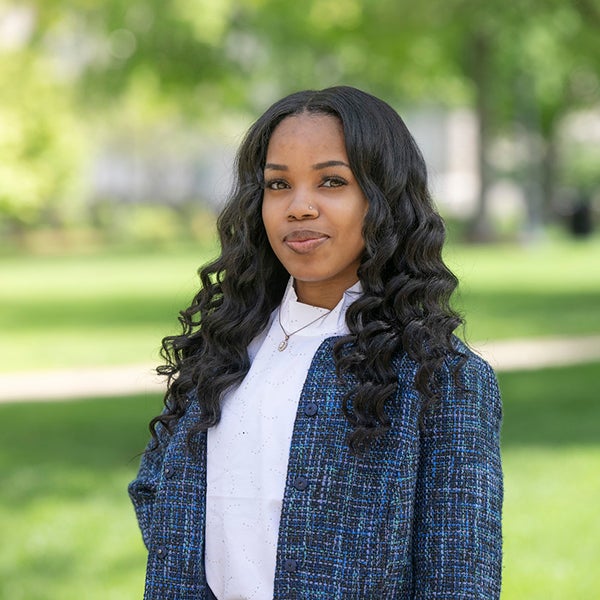 Shanisa Stinson stands smiling outdoors in a campus setting. She has long, curly black hair and is wearing a white shirt and a blue textured jacket. The background features green grass, trees, and blurred foliage.