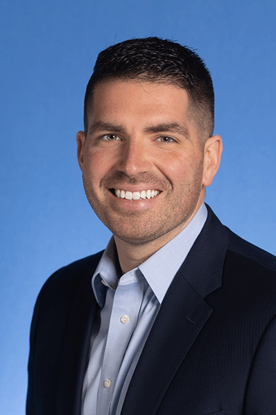 A smiling man with short, black, neatly styled hair and a goatee is wearing a dark suit jacket over a light blue dress shirt. He stands against a solid blue background. The person's hair is short and neatly styled.