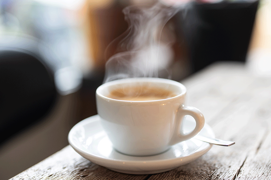 A white ceramic cup filled with steaming hot coffee sits on a matching saucer with a small spoon placed on the right side. The cup and saucer are positioned on a rustic wooden table, and the background is blurred, suggesting an indoor setting.