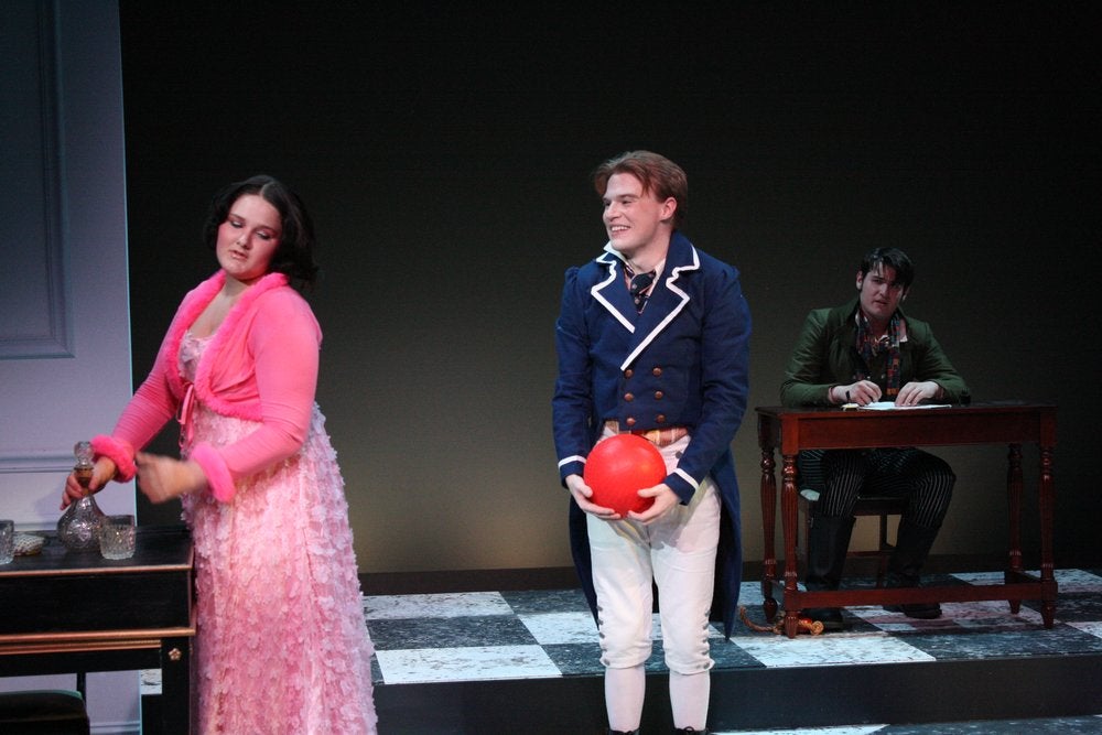 Three actors perform on stage. A woman in a pink dress stands by a table, a man in a blue coat holds a red ball, and another man in a green coat sits at a desk behind them.