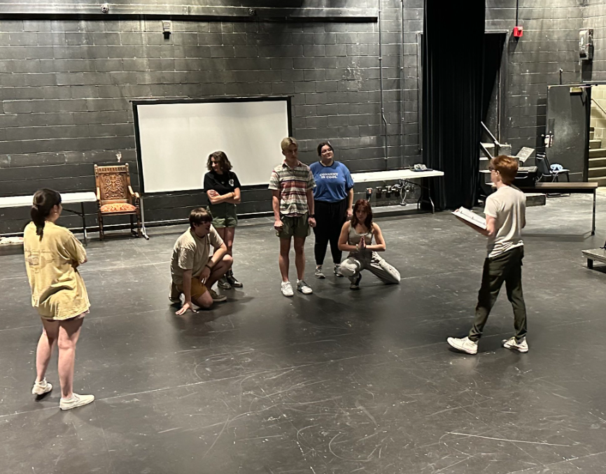 A group of seven students rehearse on a black-box theater stage. One person holding a script stands in front, while the others stand or crouch in a semicircle listening. A chair, table, and projector screen are visible in the background.