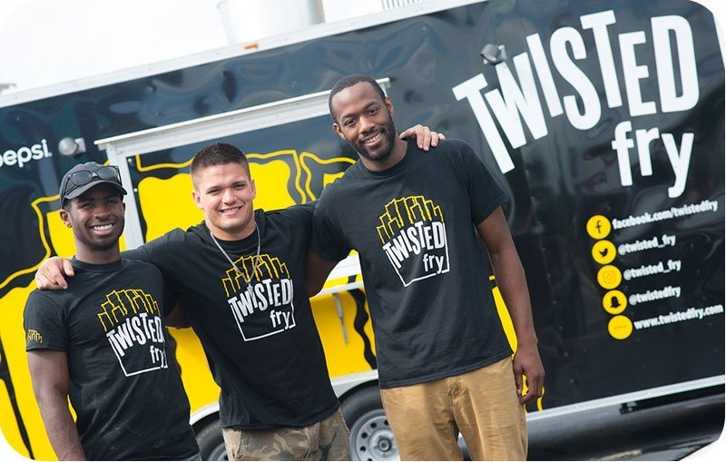 Three young men stand smiling in front of a food truck with the logo “Twisted Fry.” They are wearing matching black “Twisted Fry” T-shirts, with the truck’s black and yellow branding visible behind them.