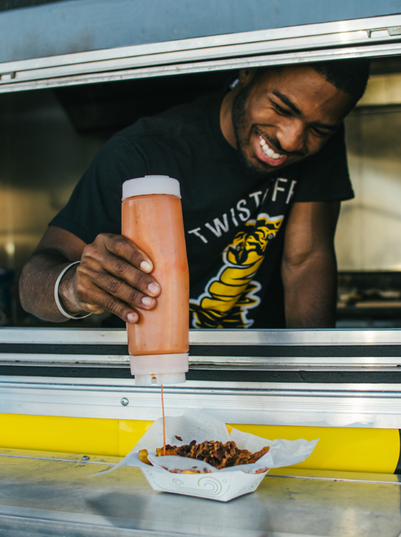 A young Black man wearing a black “Twisted Fry” T-shirt smiles while squeezing sauce from a bottle onto a tray of fries inside a food truck.