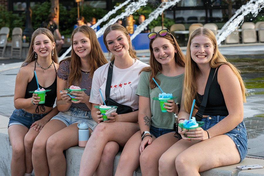 Five smiling young women sit on a ledge in front of a fountain, each holding a colorful drink with a straw. The group is casually dressed and seated close together. Behind them are chairs and green plants, suggesting an outdoor or semi-outdoor social setting.
