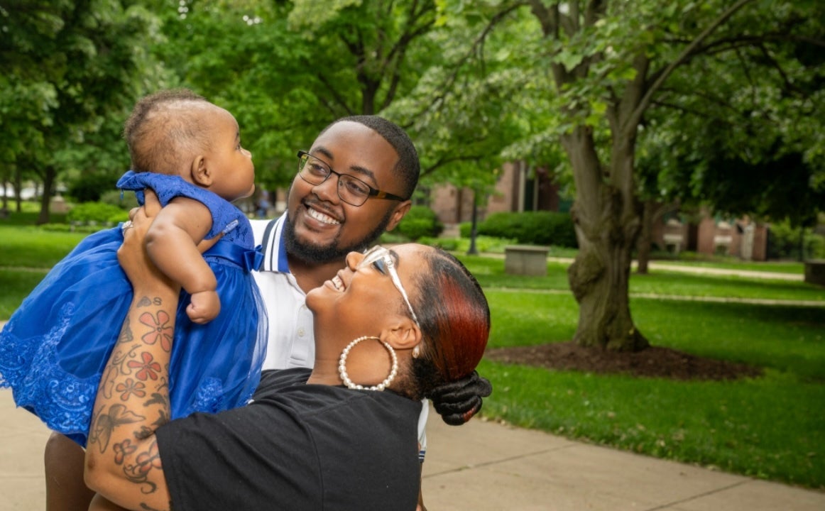 A smiling Black couple stands outdoors holding their baby dressed in a bright blue dress. The woman, wearing a black T-shirt, large hoop earrings, and clear glasses, lifts the baby toward her face, while the man, in a white collared shirt with blue trim and black glasses, looks on with a joyful expression. Green trees and walkways appear in the background.