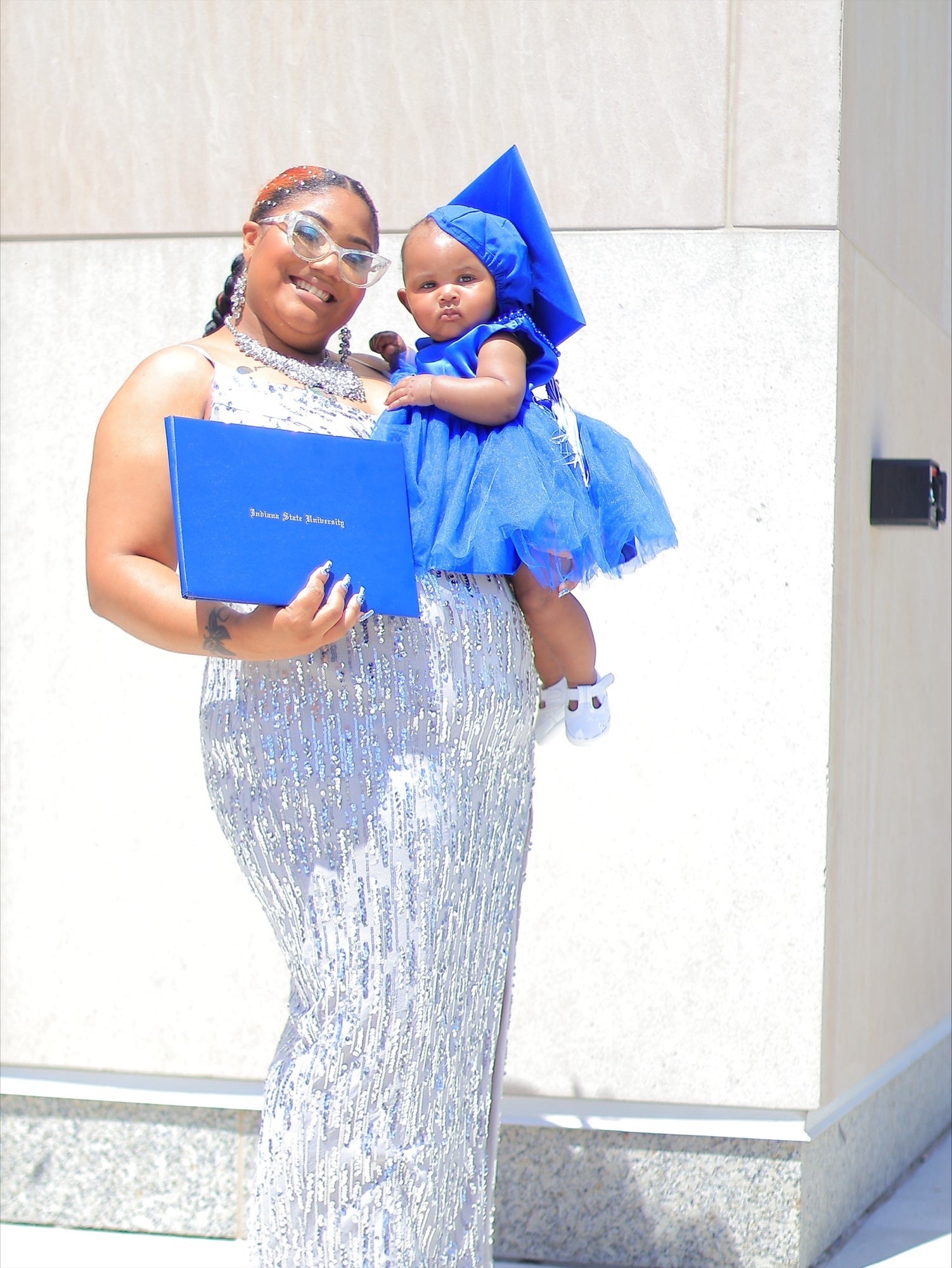 A smiling Black woman wearing a silver gown, pearl necklace, and clear cat-eye glasses holds a baby in a bright blue tulle dress and matching graduation cap. She carries a blue diploma folder reading “Indiana State University” while standing against a light-colored wall.