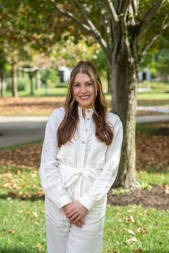 A White woman with long brown wavy hair, wearing a white long-sleeved button-up jumpsuit with a tied waist, stands outdoors smiling in a sunlit area. A tree and well-kept green grass are visible in the background.