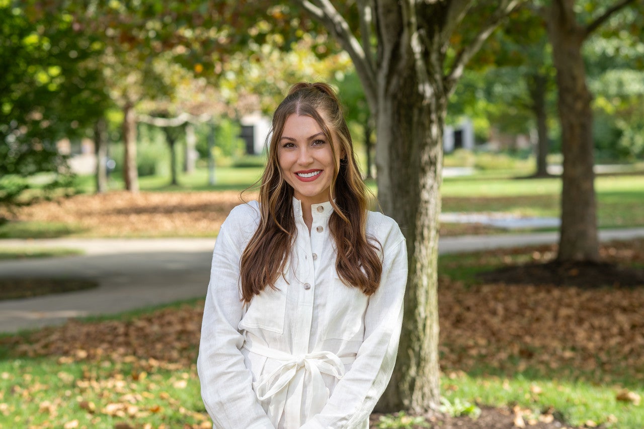 A White woman with long brown wavy hair, wearing a white long-sleeved button-up jumpsuit with a tied waist, stands outdoors smiling in a sunlit area. Trees with green and brown leaves and well-kept green grass are visible in the background.