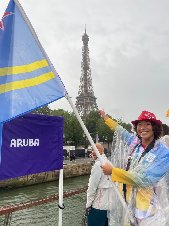 smiling young woman stands near a river with one hand pointing at the Eiffel Tower and the other holding a light blue flag with yellow stripes. She wears a red hat and a transparent rain poncho over colorful clothing, standing beside a purple banner that reads “ARUBA.”