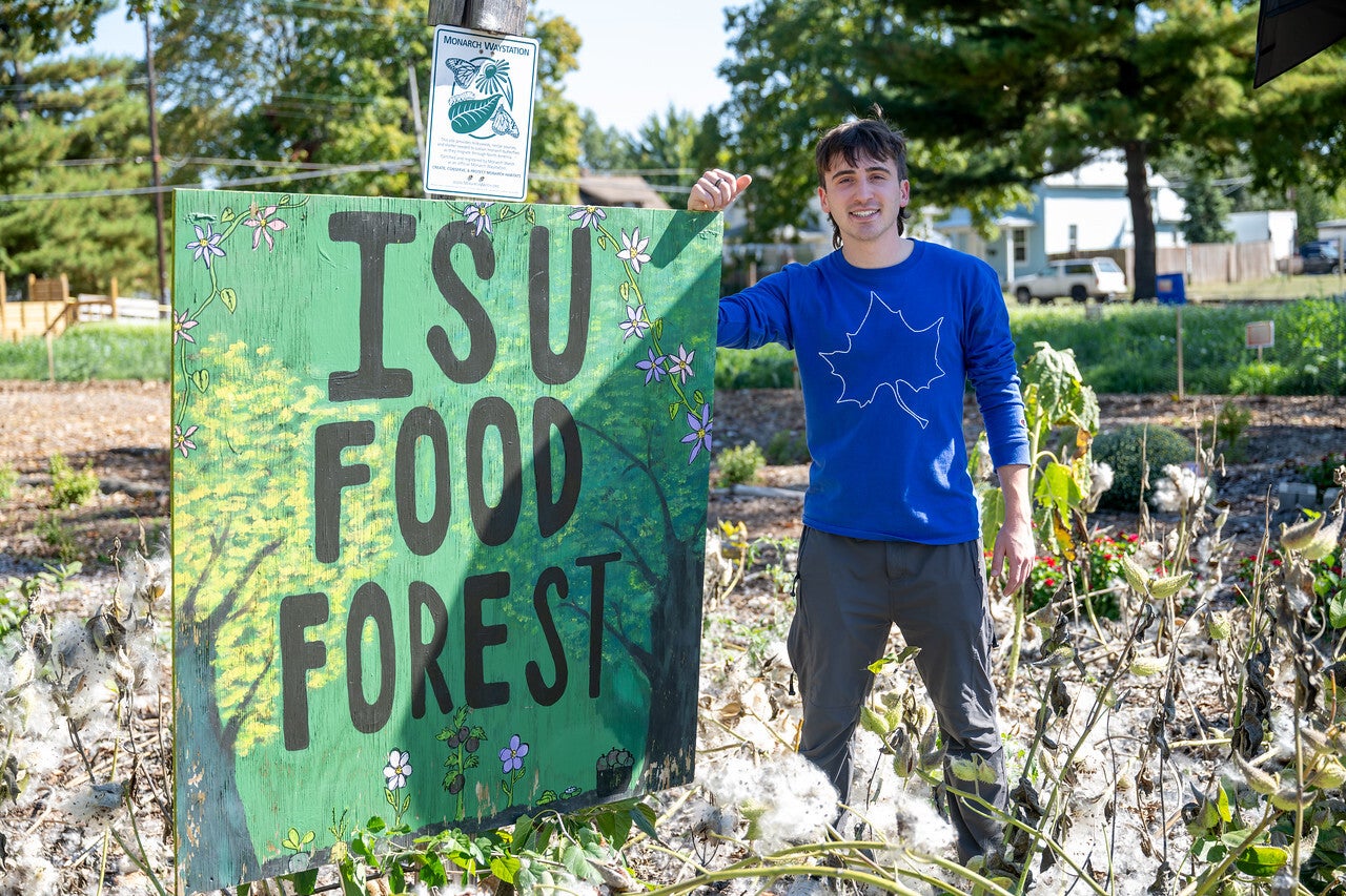 "A young man wearing a blue long-sleeve shirt with a white sycamore leaf design stands smiling beside a green sign that reads “ISU Food Forest” in a garden area."
