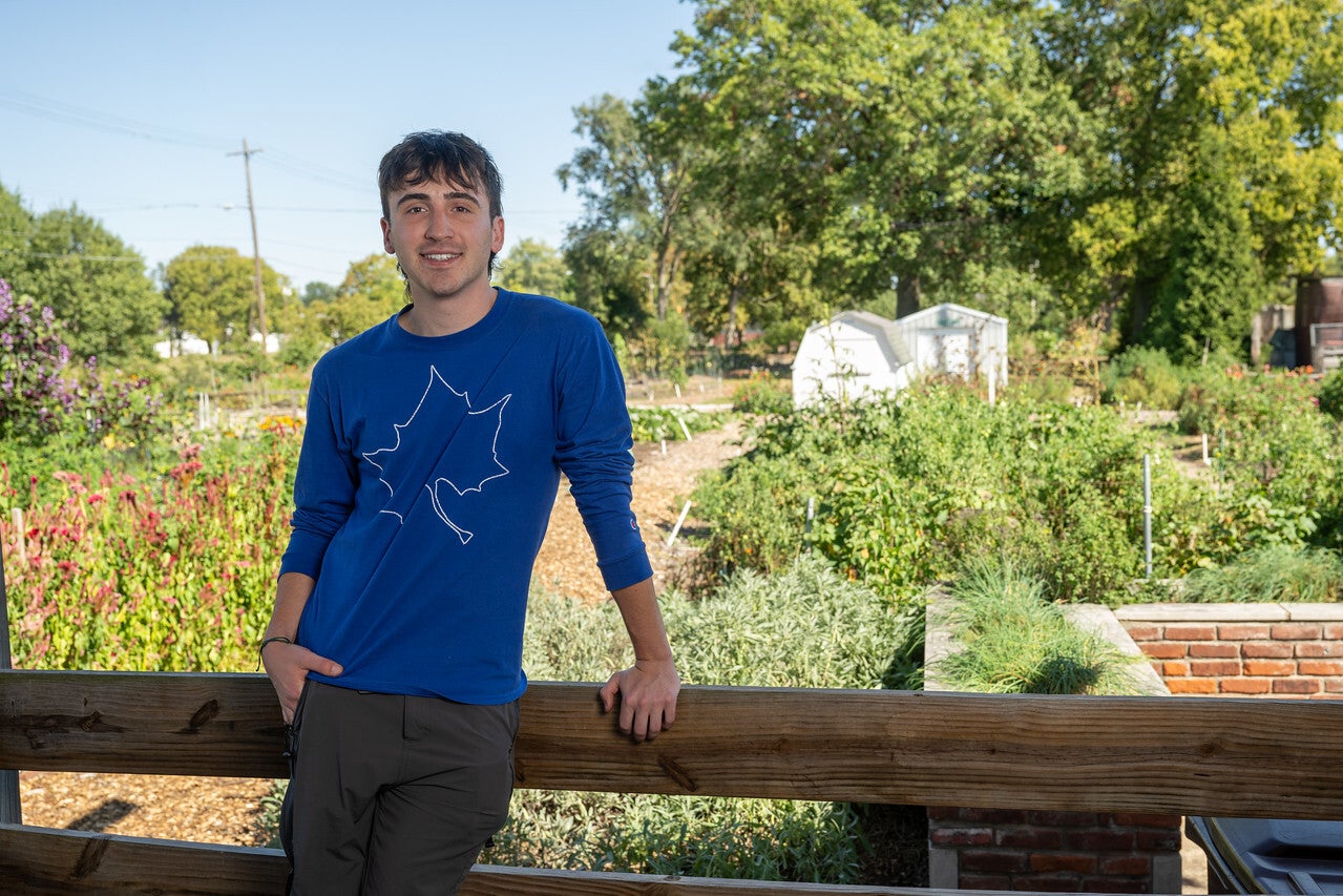 "A young White man with short brown hair wearing a blue long-sleeve shirt with a white sycamore leaf design leans against a wooden fence, smiling in a garden with plants and trees in the background."